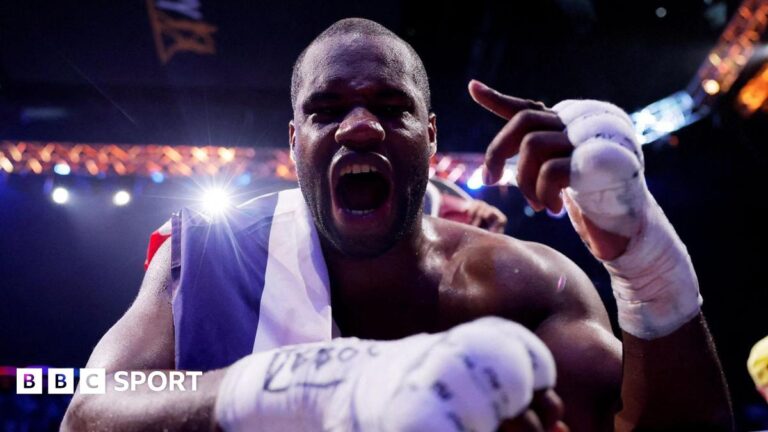 Daniel Dubois leans over the ropes of a boxing ring and holds up his hand